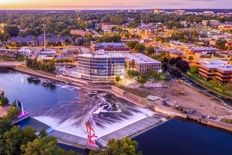 An aerial view of a downtown river with a dam and waterfall feature. Buildings with lights on line the river and city blocks. 