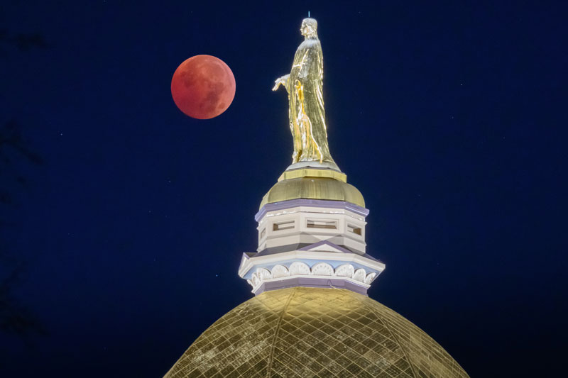 Red total lunar eclipse behind the gold Our Lady statue atop the Main Building dome at night.