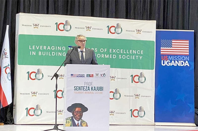 A man in a suit speaks at a lectern with the words &lsquo;US MISSION UGANDA&rsquo;on a banner behind him. 