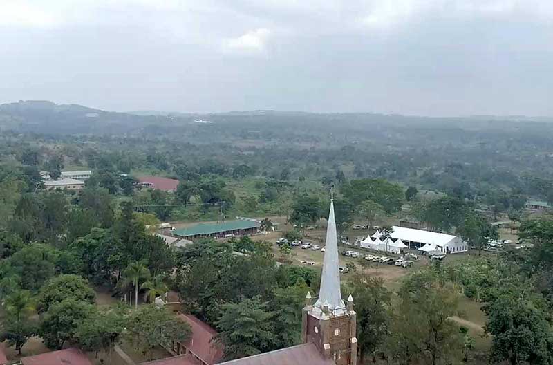 An aerial view of a landscape with a church steeple in the foreground and a large green area with several buildings and hills in the distance. 