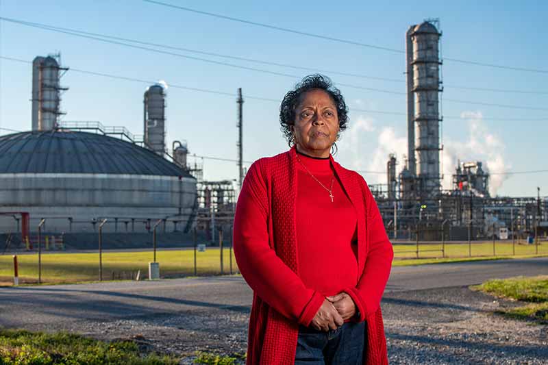 A woman in a red sweater stands outdoors, with a large industrial facility with tall smokestacks and storage tanks behind her.
