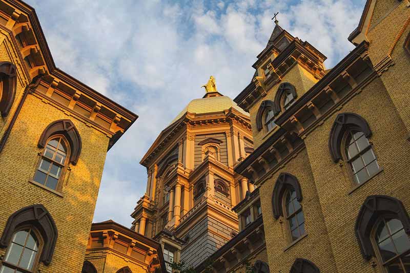A low-angle shot of the Main Building on the Notre Dame campus, showing the Golden Dome and surrounding brick architecture. 