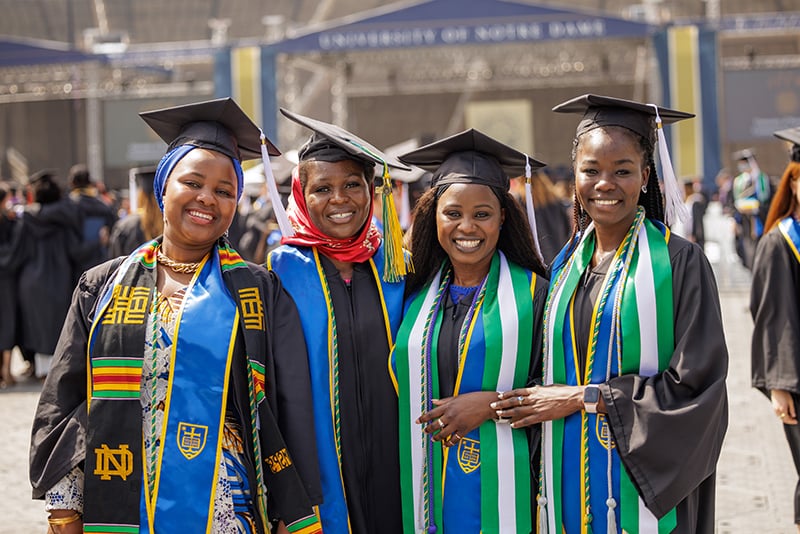 Four smiling graduates wearing black graduation gowns and colorful stoles pose for a photo. They wear mortarboards with white tassels.