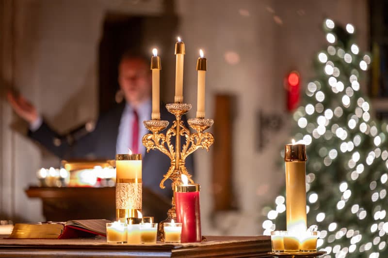 Lit candles on an altar, with an ornate gold candelabra, open Bible, and a Christmas tree in the background. A person in a suit with a red tie gestures with their hand, out of focus.