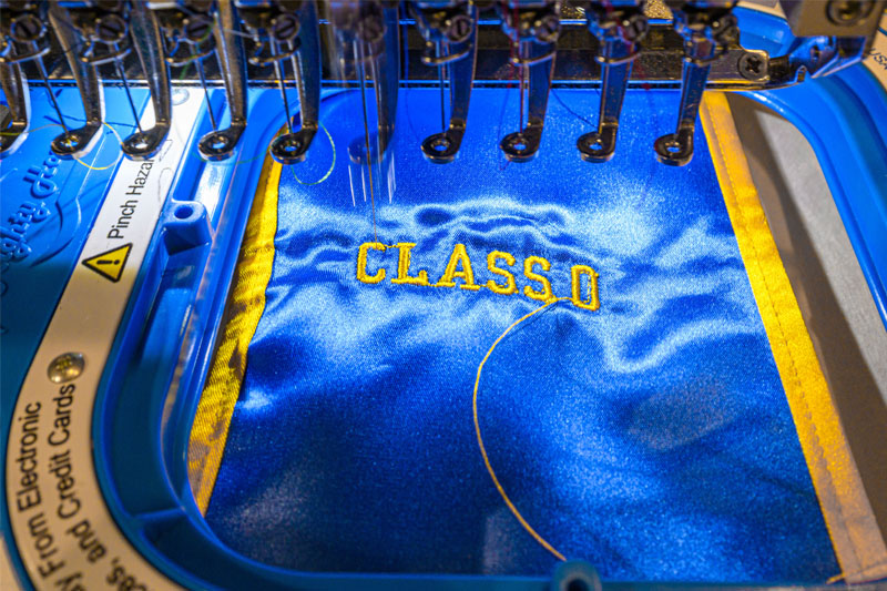 A close-up of an industrial embroidery machine at work. The machine is stitching the words CLASS OF in yellow thread onto a piece of blue satin-like fabric.