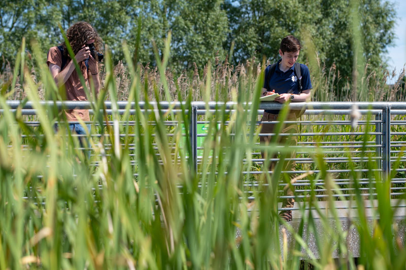 Two students, one male and one female, standing on a bridge or walkway in a natural, reedy environment. The person on the left is holding a camera, while the person on the right is writing in a notebook.