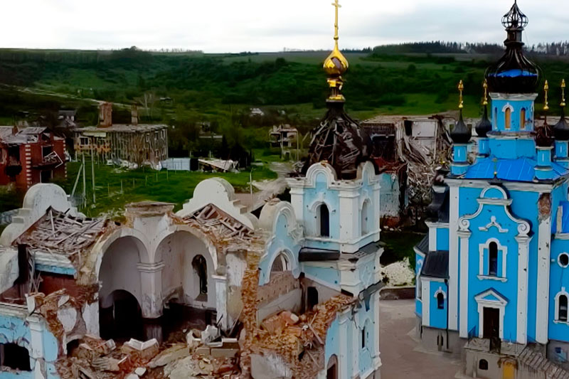 An aerial view of two churches, one blue and white, the other white and light blue, standing amongst the ruins of other buildings. The white and light blue church is heavily damaged with its roof caved in and rubble surrounding its base. The blue and white church appears intact with a bright blue tarp covering sections of its roof.