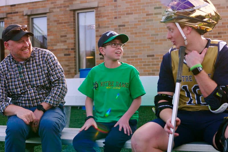 A man in a plaid shirt and a boy in a green Notre Dame shirt sit on a bench and talk to a Notre Dame lacrosse player in uniform.
