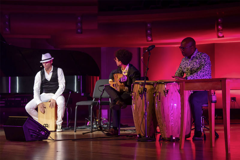 Three musicians perform on a stage bathed in pink light. One, wearing a white fedora and black vest, plays a cajón. Another plays an acoustic guitar. The third sits at a table playing congas. A grand piano and other equipment are visible on stage.