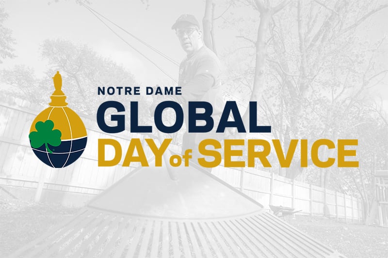 A graphic for the Notre Dame Global Day of Service. The logo, which features a globe with a shamrock and the statue of Mary from the Golden Dome, is prominently displayed to the left of the text. The background is a semi-transparent black and white photo of a person using a large leaf rake in a yard with a wooden fence.