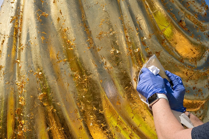 A close-up of a person's hands wearing blue gloves, applying gold leaf to the surface of the Golden Dome.