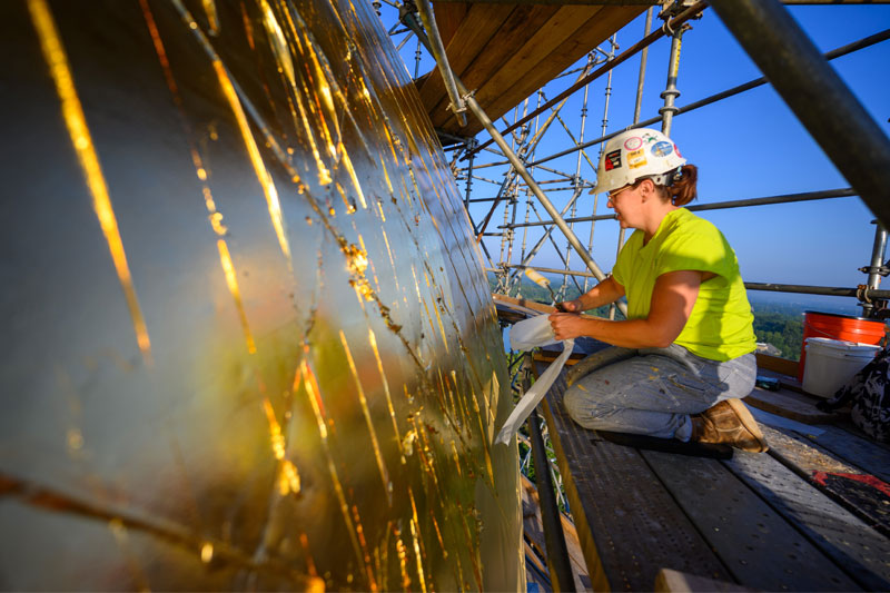 A female kneeling on a scaffold, applying gold leaf to the surface of the Golden Dome. They are wearing a hard hat, a bright yellow t-shirt, and paint-splattered jeans.