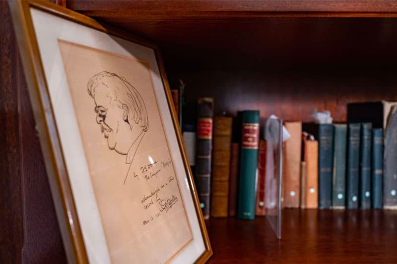 Framed caricature sketch of a man with a mustache, facing left, signed and dated March 18, 1954, leaning against books on a wooden shelf.