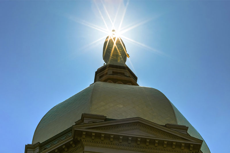 The golden dome of the Main Building at the University of Notre Dame. A prominent sunburst effect is centered on the statue of the Blessed Mother atop the dome, with a clear blue sky in the background.