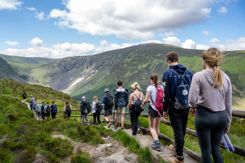 Students with backpacks descend a rocky, grassy path with a wooden railing, overlooking a green valley and mountains under a blue sky with clouds.