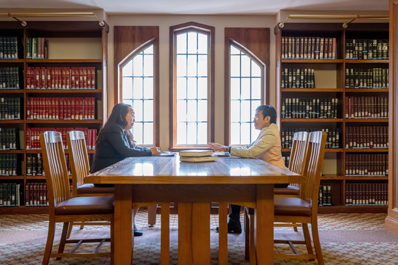 Two people sit at a wooden table between bookshelves, conversing near a bright window.