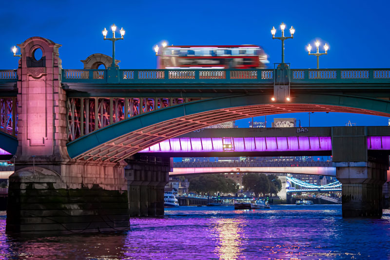 A blurred red double-decker bus crosses a teal metal bridge at twilight. The bridge's underside is illuminated with purple light, and the Tower Bridge is visible in the distance downstream. Riverboats and a city skyline are also visible beyond the bridge.