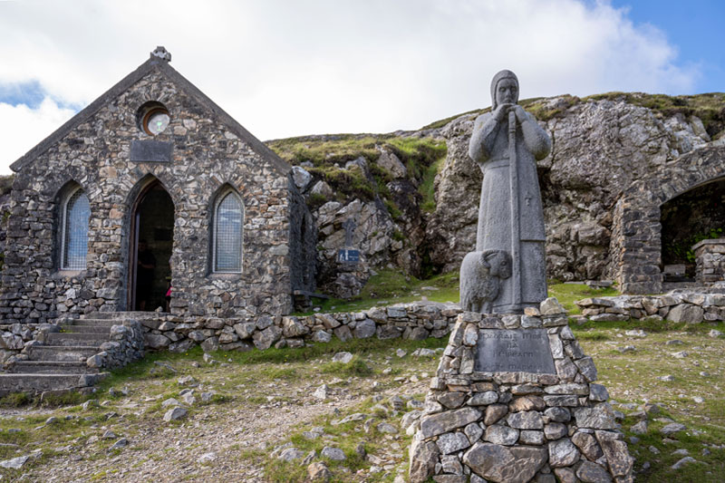 Stone statue of a robed figure with a staff and lamb, beside a small stone church built into a hillside.