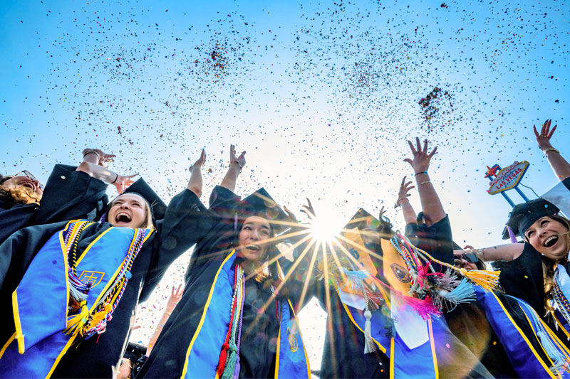 A group of student throwing confetti in the air. The sun is shining brightly in the background.