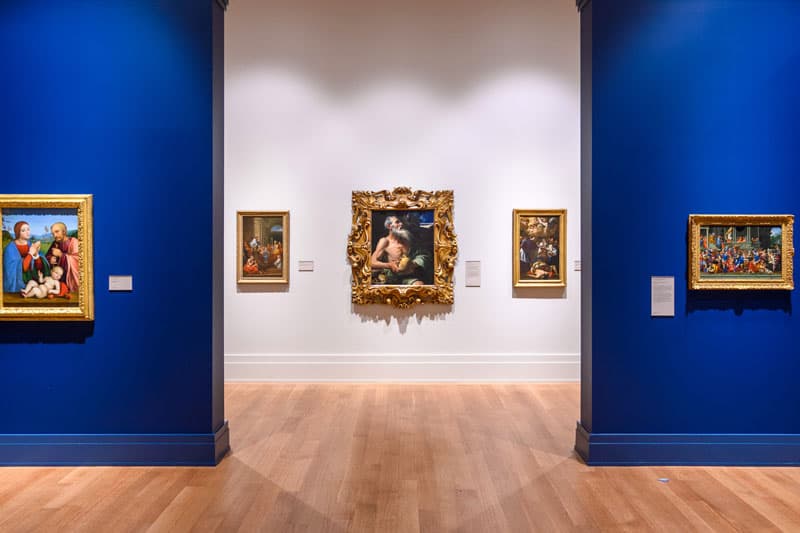 Gallery view in the Snite Museum of Art at the University of Notre Dame, with framed religious paintings on royal blue and white walls. Light wood floors reflect the artwork.