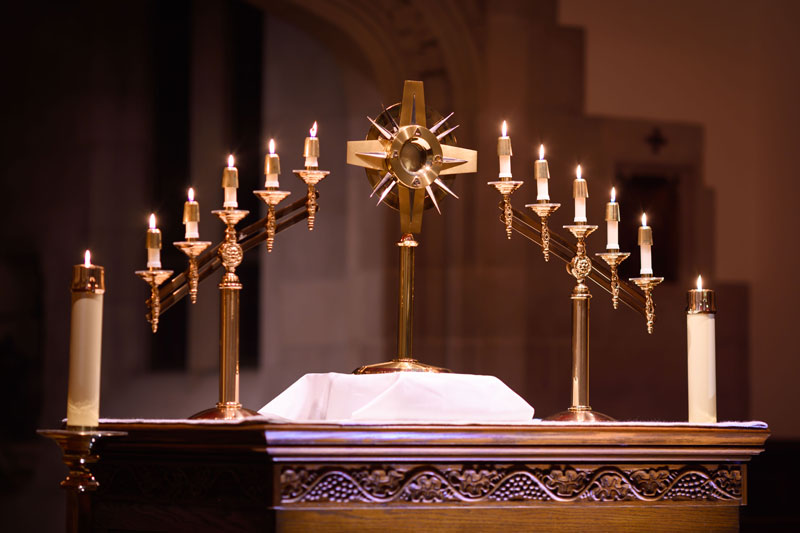 A monstrance, a vessel used to display the consecrated Eucharist, stands on a wooden altar. Lit candles on ornate brass candelabras flank the monstrance, illuminating the altar in a dimly lit church. Two larger candles sit on the edge of the altar on either side.