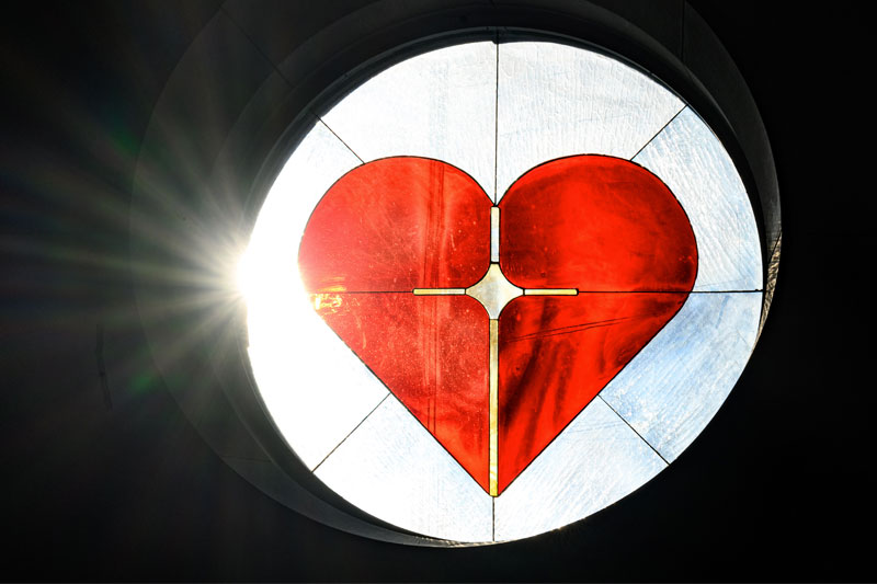 Bright sunlight shines through a circular stained-glass window depicting a red heart with a cross superimposed.