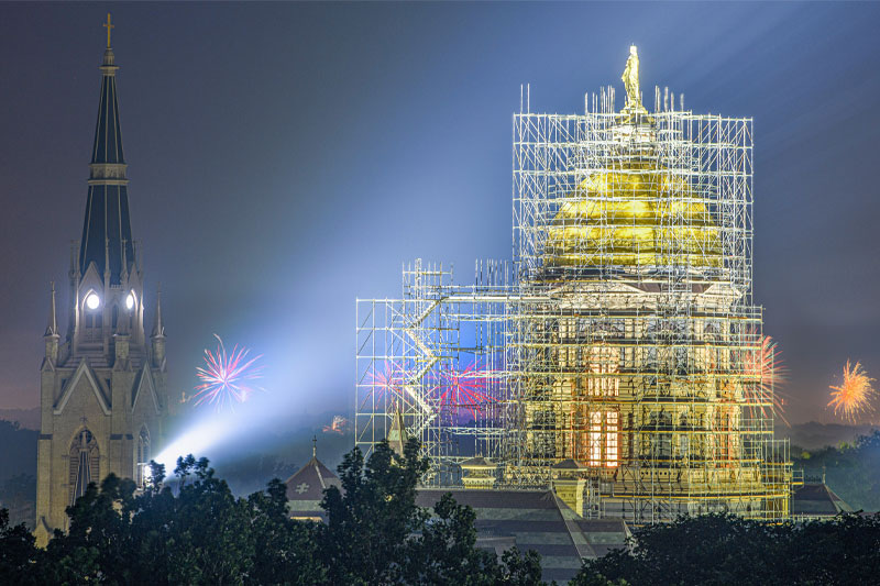 A night-time view of two prominent buildings. On the left, there is a tall church steeple with a clock face and a glowing light near the top. On the right, a large golden dome is surrounded by extensive scaffolding. Atop the Golden Dome is a statue. Fireworks are visible in the background.