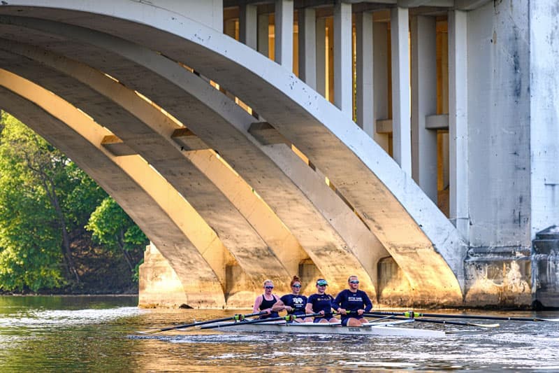 Four Notre Dame rowers in a scull passing beneath the white arches of a bridge.