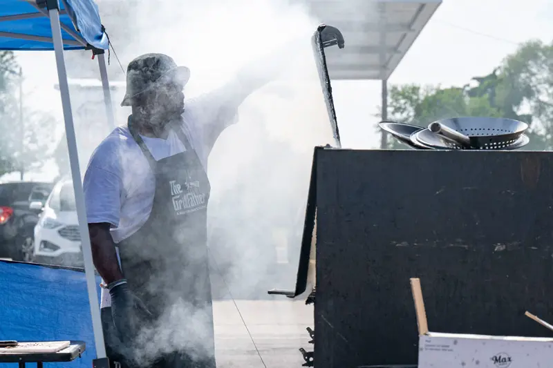 A black man stands in front of his port-a-pit grill while smoke envelopes his face.