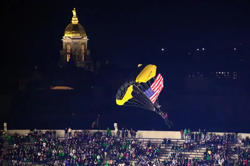 At night, a parachutist with a black and gold parachute descends into Notre Dame Stadium, carrying an American flag. The illuminated Golden Dome of the Main Building is visible in the background, while spectators in the stands watch.
