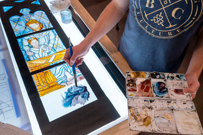 An artist paints a section of a stained-glass window depicting Saint Joseph and Jesus using a blue hue from a watercolor palette on a light table.