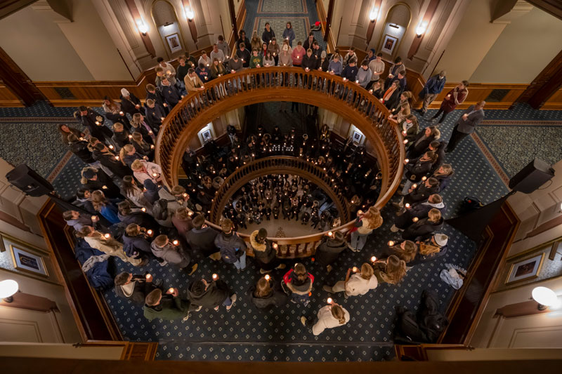 Students hold lit candles on the balconies and grand staircase of the Main Building at the University of Notre Dame. The view is from above, looking down the spiral staircase.
