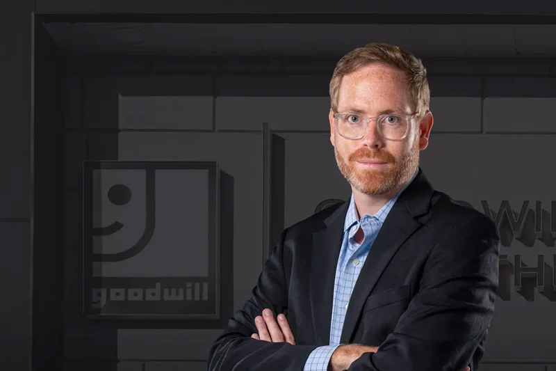 A man with glasses and a red beard wearing a blazer and light blue collared shirt stands with his arms crossed in front of a gray wall featuring several Goodwill logos.