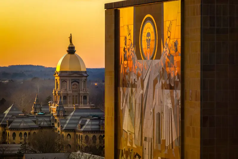 At sunset, the golden dome of the Main Building at the University of Notre Dame is juxtaposed with a segment of the iconic Word of Life mural known as Touchdown Jesus.