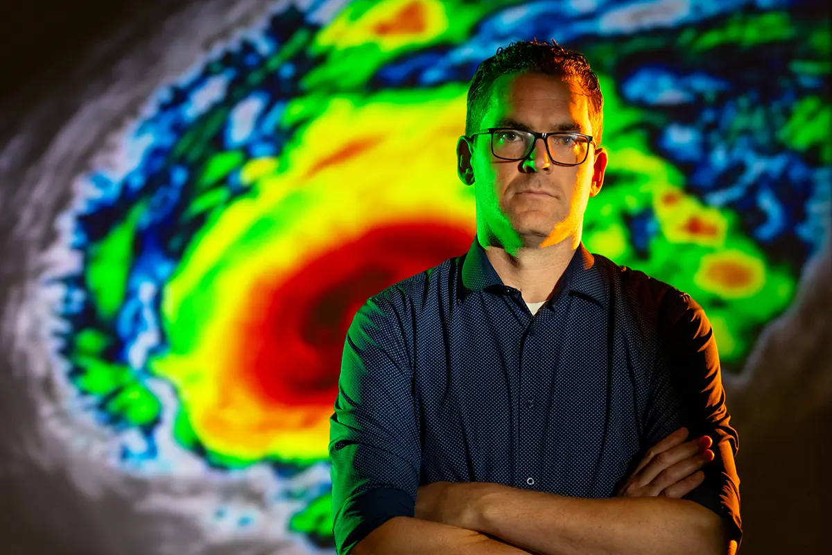 A serious man in a dark blue shirt and glasses stands with his arms crossed in front of a colorful hurricane projection.