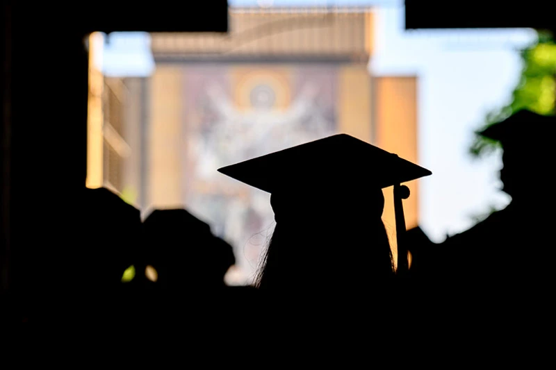 Silhouetted graduates in caps and gowns face the Hesburgh Library's &ldquo;Word of Life&rdquo; mural at Notre Dame, seen through an archway.