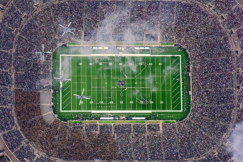 Aerial view of Notre Dame Stadium filled with fans during a performance. Four helicopters fly over the green field where the Band of the Fighting Irish forms the ND logo.