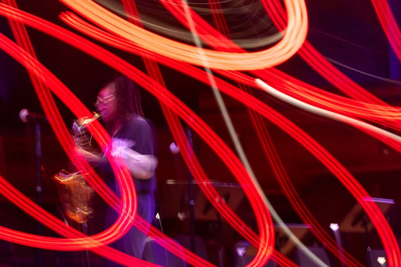 A saxophonist plays on a dark stage. Blurred streaks of red and white light from stage lighting crisscross in front of them. The lights create an abstract, energetic visual effect.