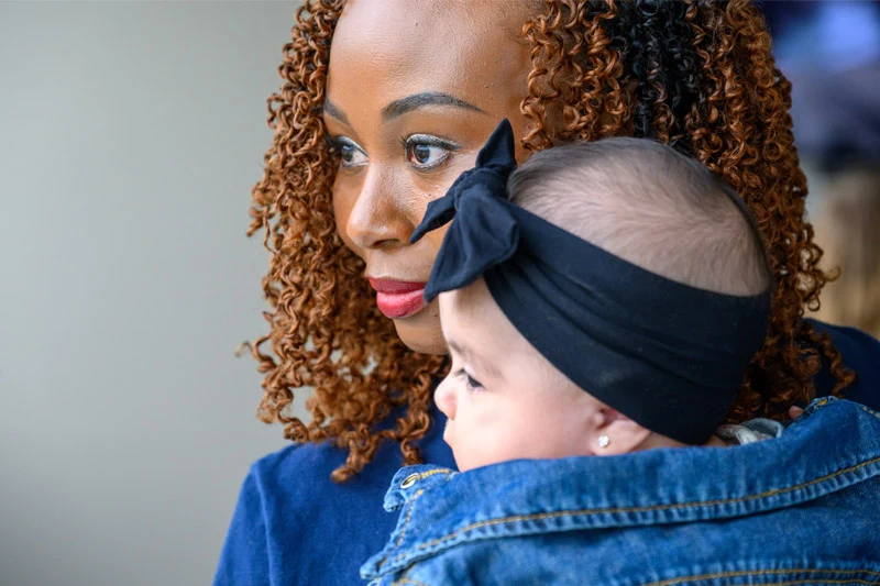 A woman with brown curly hair and red lipstick holds a baby wearing a denim jacket and black bow headband.