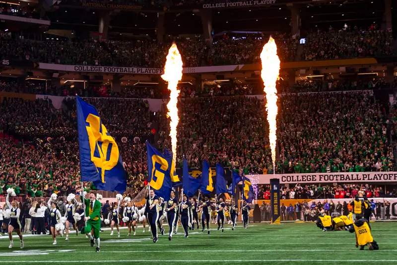 Notre Dame cheerleaders and flag holders run onto the football field as flames shoot up behind them. The stadium is packed with fans in the background, many wearing green.
