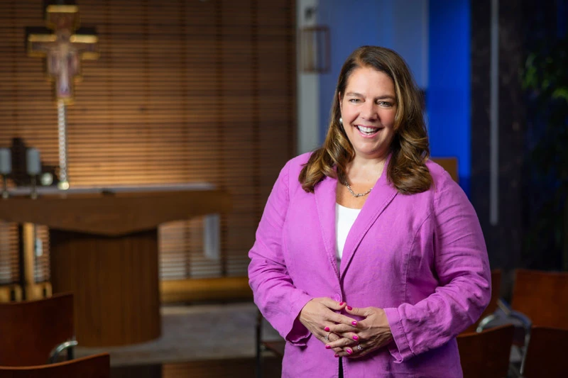 A woman with shoulder-length brown hair, wearing a fuchsia blazer and white shirt, smiles with her hands clasped in front of her in a small chapel. A crucifix and altar are visible in the background.