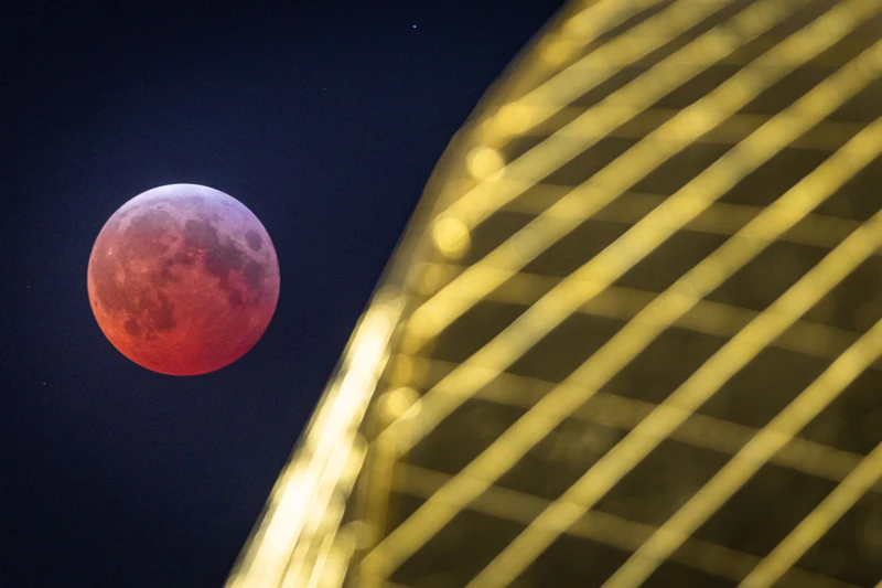 Reddish-orange moon during a lunar eclipse with a blurred, out-of-focus section of the illuminated Golden Dome at the University of Notre Dame.
