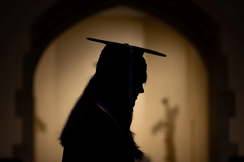 Silhouette of a graduating student wearing a cap and gown, head bowed.