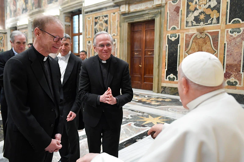 Two priests in black suits and clerical collars clasp their hands and smile as they speak with Pope Francis, who is seen from behind wearing his white zucchetto and cassock.