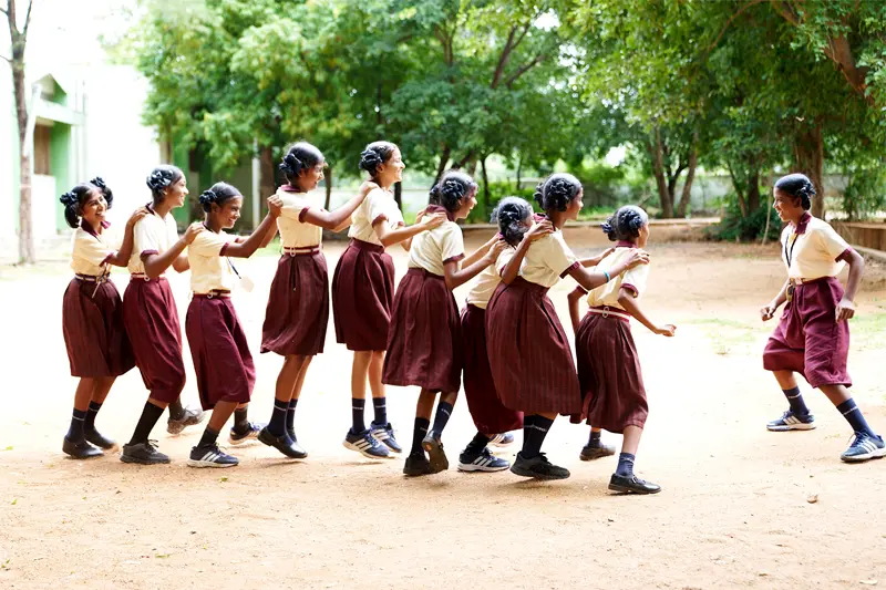 A line of Indian girls in matching tan and maroon school uniforms hold each other's shoulders and playfully skip across a dirt schoolyard. Trees and a school building can be seen in the background.
