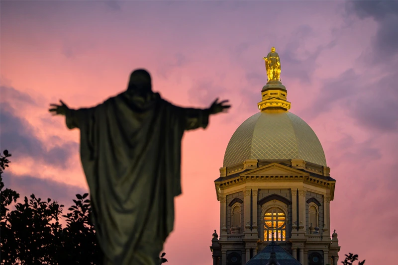 Silhouetted statue with outstretched arms against a vibrant pink and orange sunset, with the golden dome of the Main Building and its statue of Mary at the University of Notre Dame.