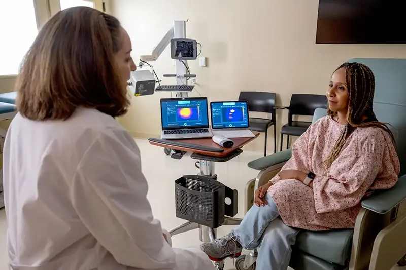 A healthcare professional in a white coat speaks with a patient in a pink hospital gown, while two laptops display medical imaging on a rolling cart.