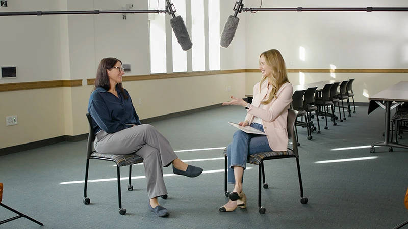 Two women sit facing each other in chairs in a large room, with microphones suspended above them for a podcast recording.