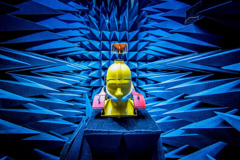 A yellow mannequin head with red and white attachments sits surrounded by blue sound-dampening foam cones in an anechoic chamber.