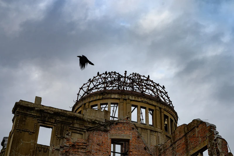 The ruined Hiroshima Peace Memorial (Genbaku Dome) featuring exposed brick and a skeletal metal dome against an overcast sky. Several dark crows perch on the dome, one flying past.